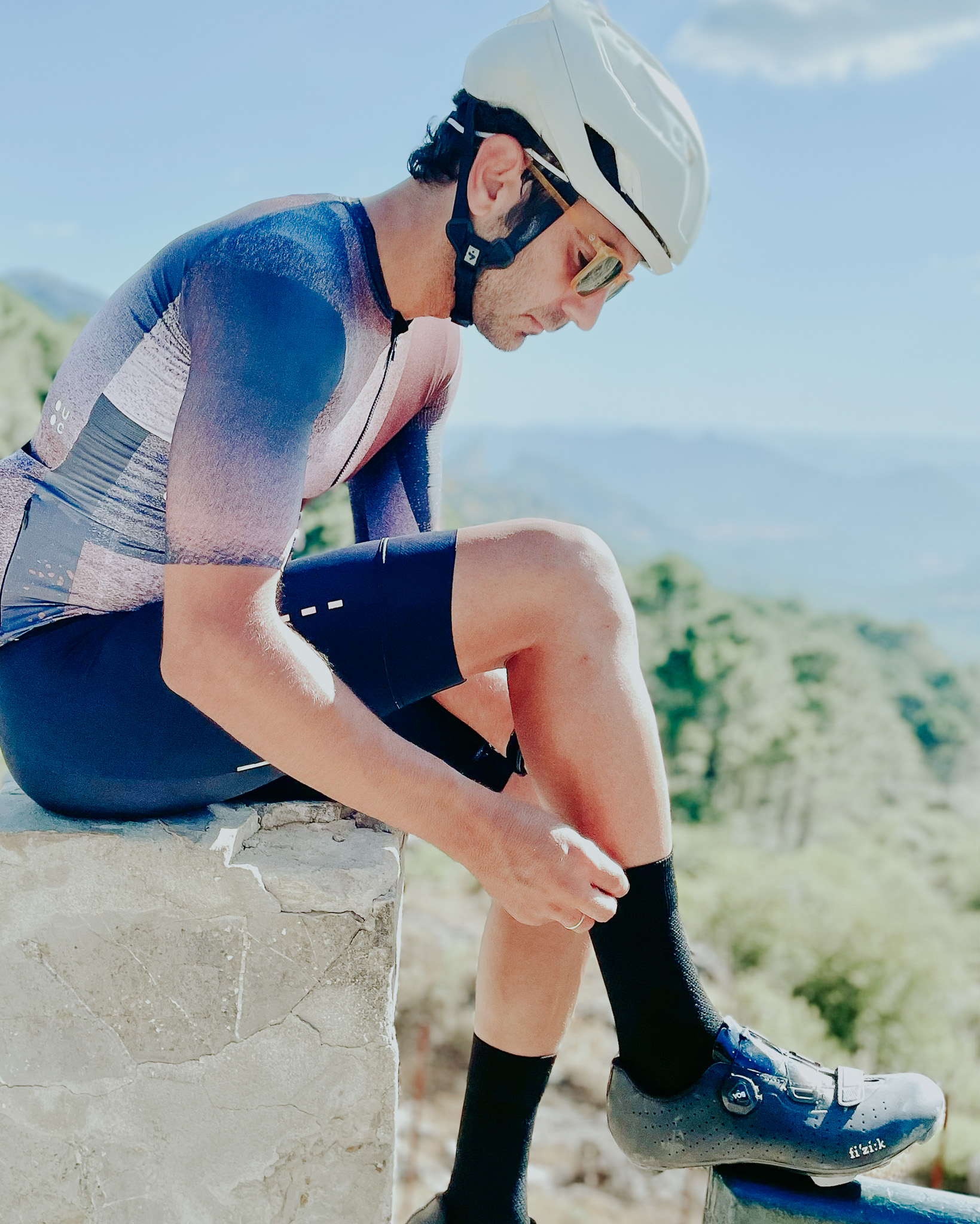 Cyclist adjusting black gravel socks while sitting on a stone ledge, wearing a white helmet, sunglasses, and a sleek cycling kit, with a scenic mountainous backdrop.