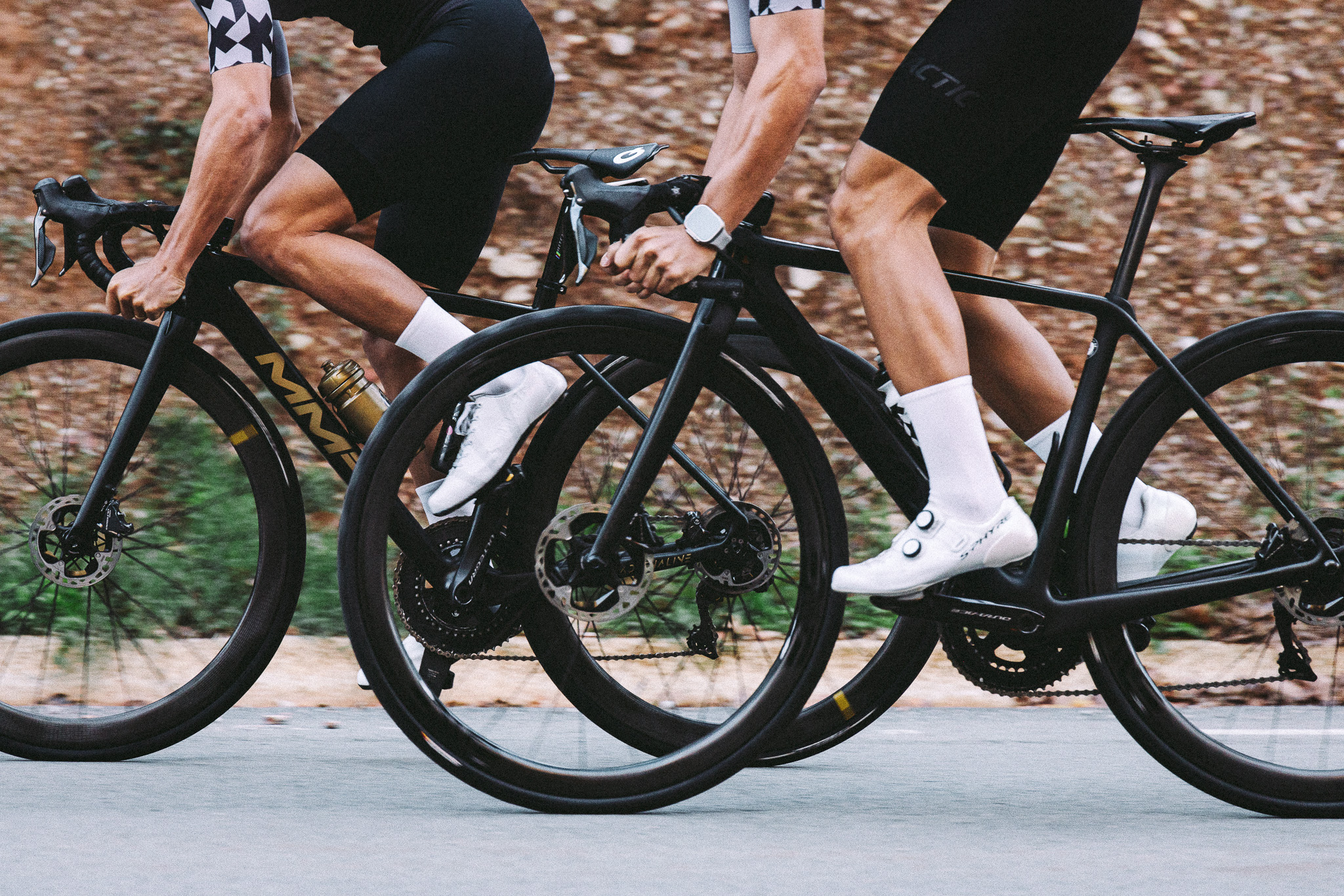 Two cyclists wearing black cycling kits and white long socks riding high-performance road bikes on a scenic outdoor trail.